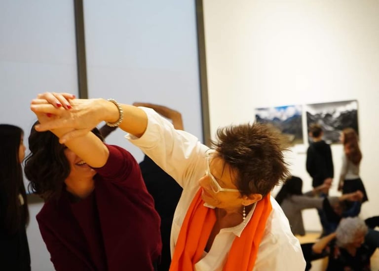 Giovane e anziana danzano insieme tenendosi per mano in museo, sullo sfondo altri danzano come loro