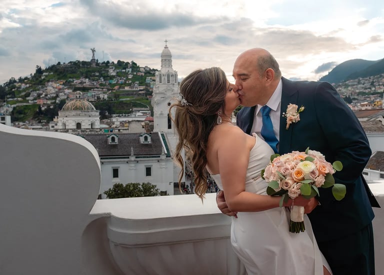 El novio besando a la  novia mientras la agarra de la cintura en un terraza con vista al Panecillo en el centro histórico 
