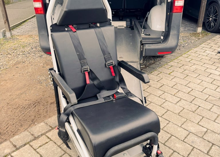 Mobilitätsstuhl mit roten Gurten auf Rampe hinter grauem Fahrzeug, draußen.