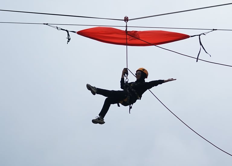 A person wearing a helmet and harness glides on a zipline under a red canopy against a cloudy sky.