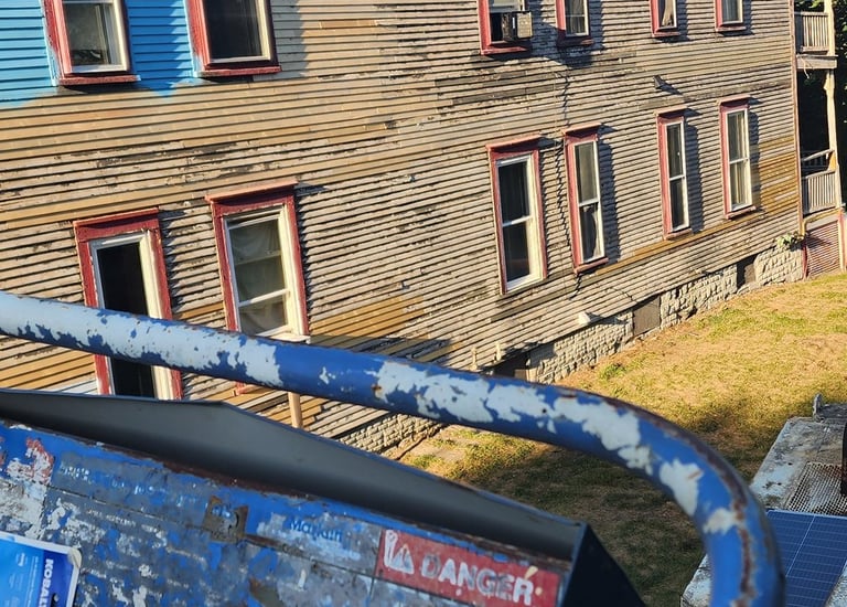 A blue scissor lift basket overlooks a residential building with weathered beige wood siding.