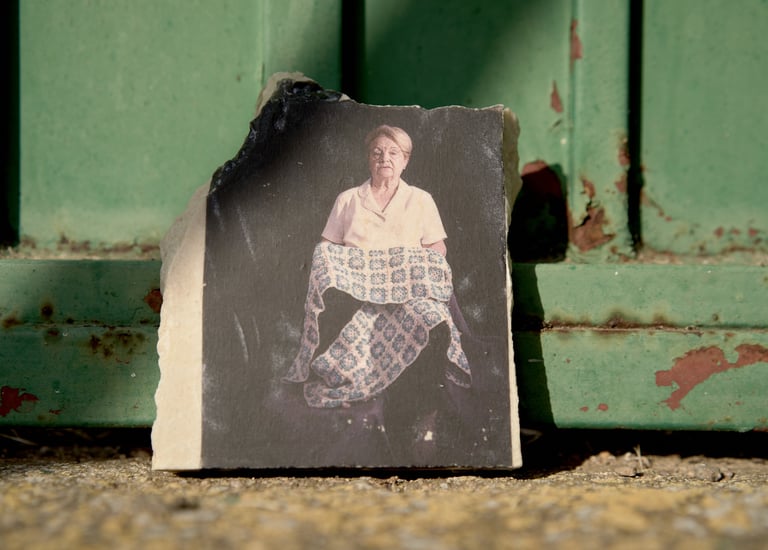 A vintage portrait of an elderly woman holding a patterned quilt on a stone fragment against a green metal door.