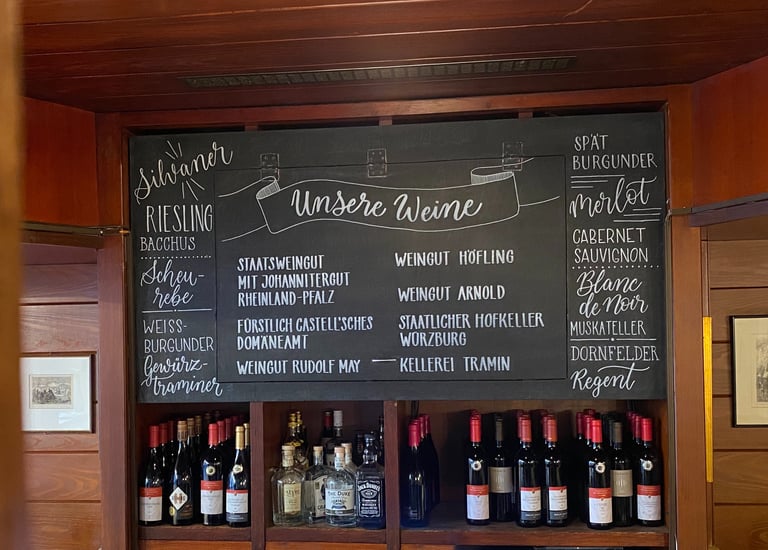 Handwritten wine menu on a chalkboard featuring German Riesling and Silvaner above shelves of wine bottles.