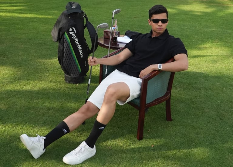 A young golfer wearing sunglasses relaxes in a chair on a green golf course with his TaylorMade bag and clubs.