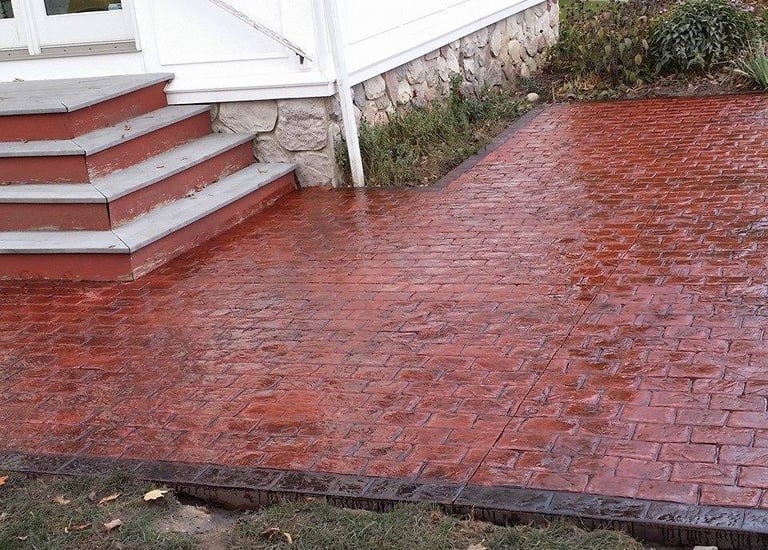 Custom red stamped concrete patio with decorative stone borders installed next to a white house.