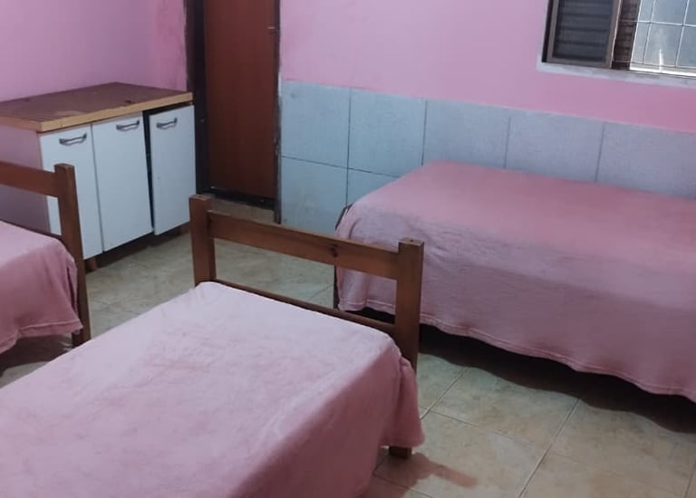 Three single beds with pink linens in a modest bedroom with pink walls and tile flooring.