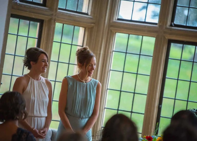 Smiling brides in elegant gowns standing by a large stone window during their wedding ceremony.