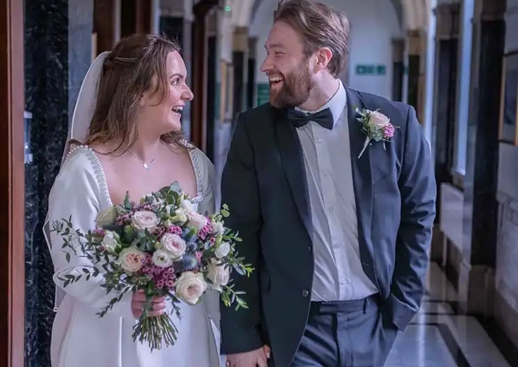 Smiling bride in white gown and groom in tuxedo walking down hallway with a floral bouquet.