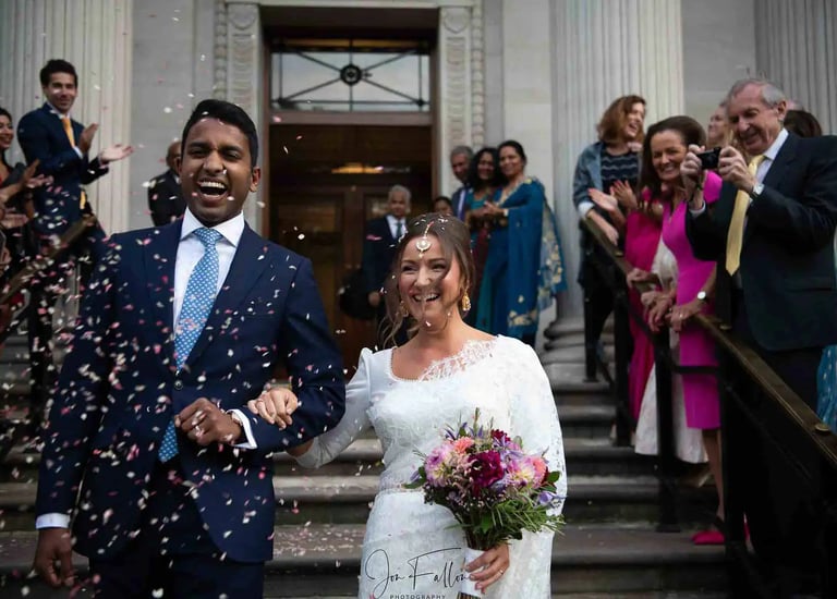 Smiling bride and groom walk through falling confetti after their wedding ceremony at a historic building.