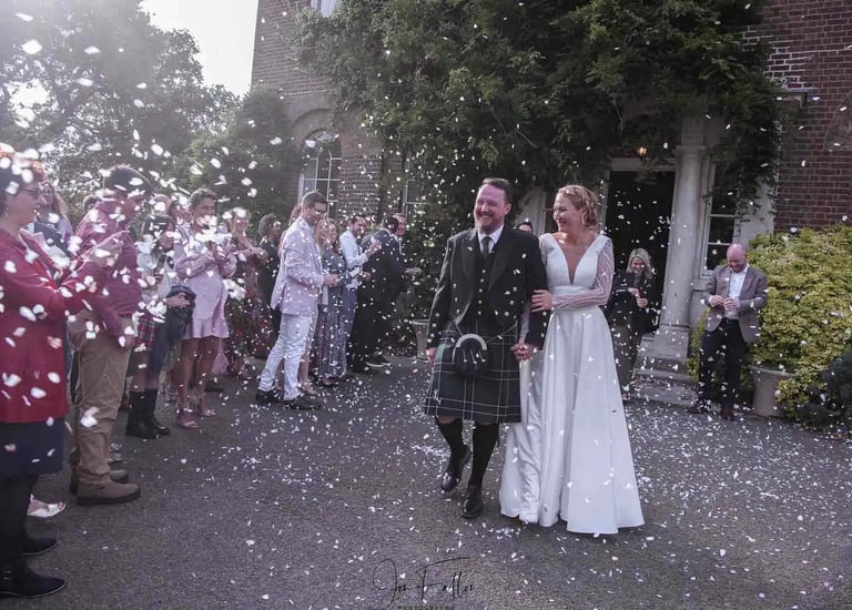 A bride and groom in a traditional kilt walk through falling confetti at an outdoor wedding.