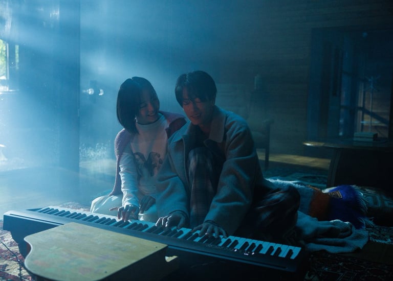 A man and woman play a digital piano keyboard together in a misty, blue-lit room.
