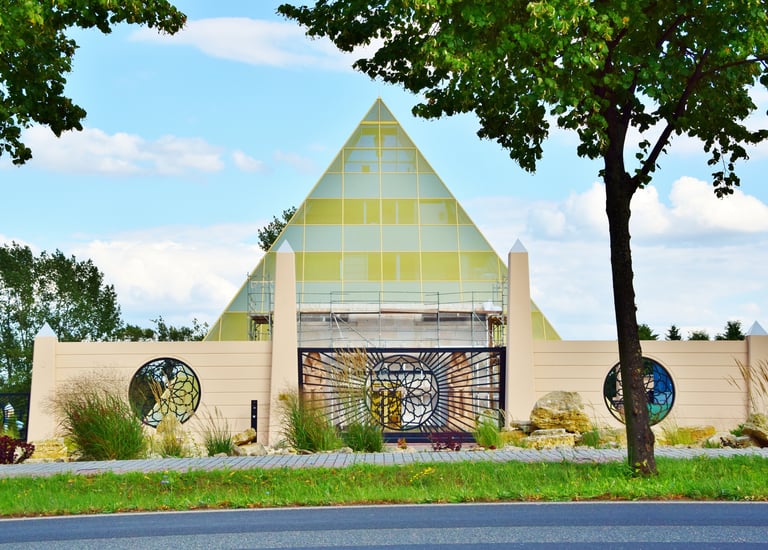 Modern pyramid-shaped building with a sunburst gate and circular windows framed by lush green trees.