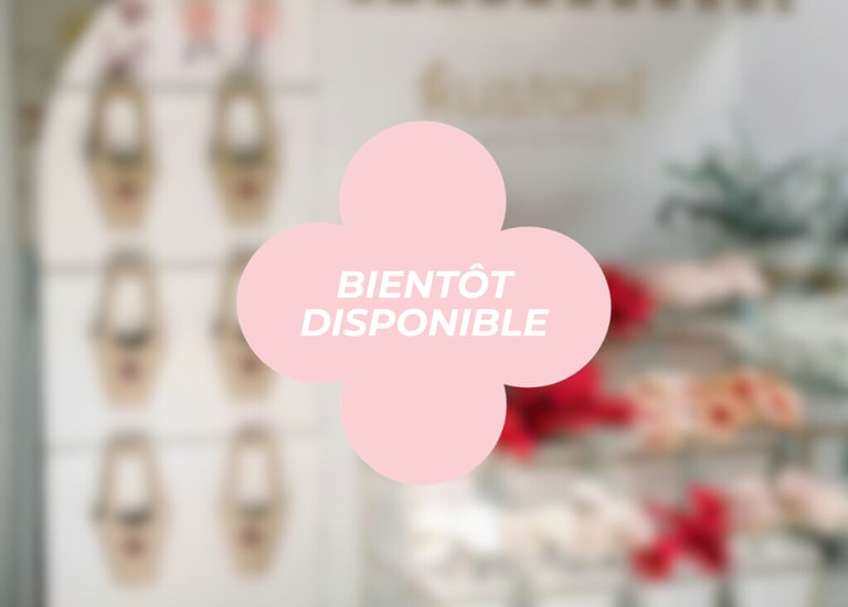 Pink flower-shaped 'Bientôt Disponible' coming soon sign overlaying a blurred boutique flower shop interior.