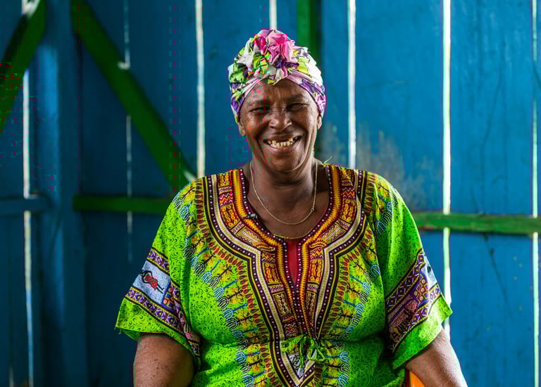 Femme souriante vêtue d'un dashiki vert vif et d'un foulard floral coloré, sur fond bleu bois.