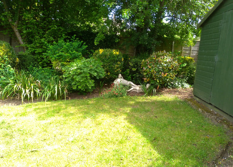 Backyard garden with green lawn, lush bushes, and stone statues next to a green shed.