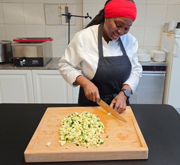 Chef Viviane in white chef jacket and black apron chopping vegetables on a bamboo board in a professional kitchen