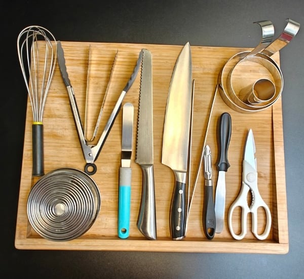 Professional chef tools neatly arranged on a bamboo board including a whisk, knives, tongs, pastry rings and kitchen scissors