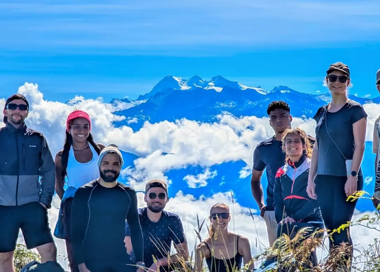 Diverse group of hikers posing on a mountain summit above clouds with snow-capped peaks in the background.