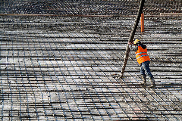 Worker with orange vest and yellow helmet holding boom pump hose for concrete covering wire mesh.