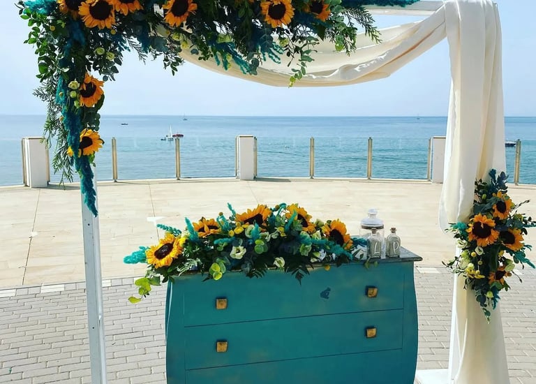 Seaside wedding ceremony setup featuring white arch and teal vintage dresser, adorned with sunflower and turquoise foliage