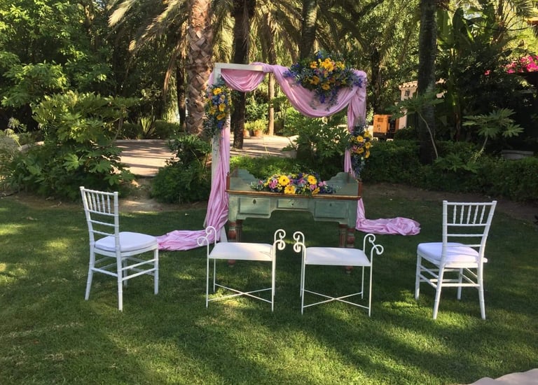 Outdoor wedding ceremony setup in lush garden, pink fabric-draped arch floral arrangements, vintage green desk, white chairs