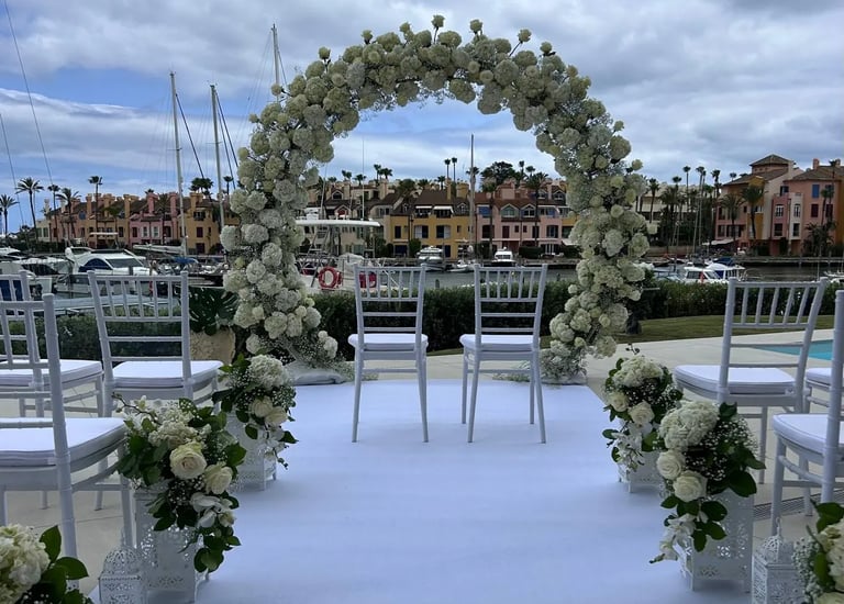 Elegant outdoor wedding ceremony setup featuring a large circular white floral arch, white chairs overlooking a marina.