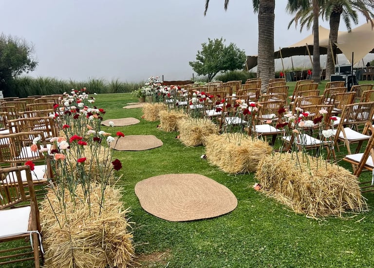 Outdoor wedding ceremony seating with wooden chairs, jute rugs, and colorful carnations growing from hay bales.