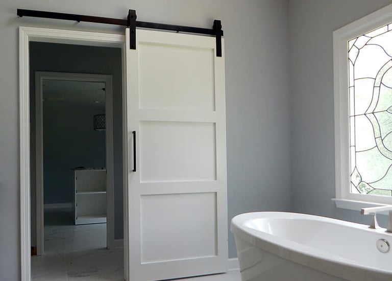 White sliding barn door with black hardware installed in a modern bathroom near a soaking tub.