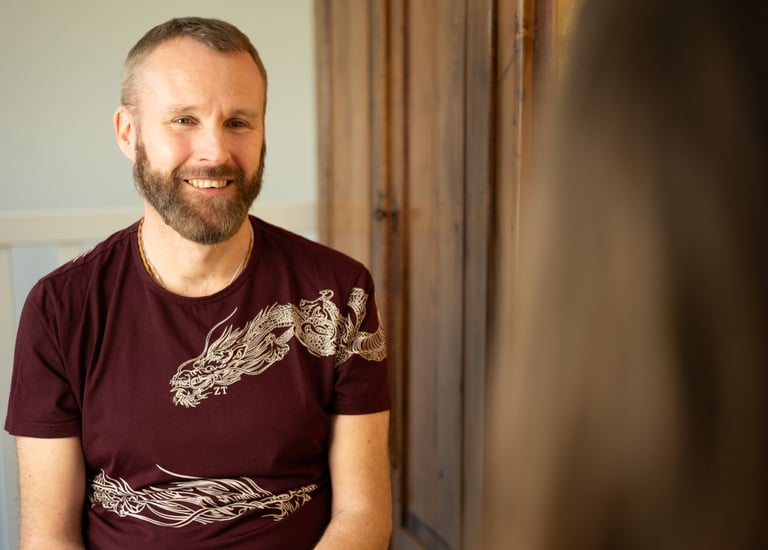 A man wearing a dark red t shirt with dragon imagery is sitting coaching a client