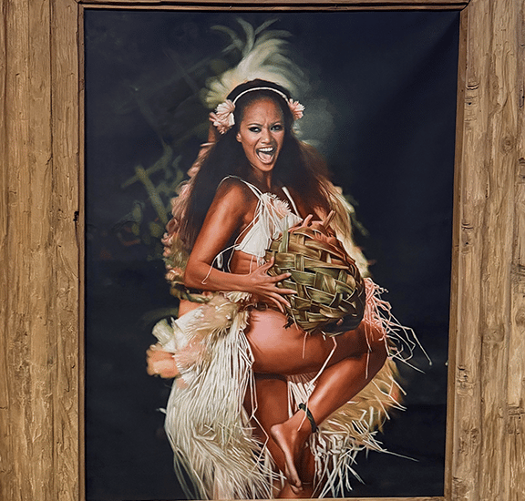 Polynesian hula dancer in traditional grass skirt performing in a rustic wooden frame portrait.