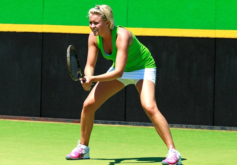 A blonde female tennis player in a green top and white skirt waits to return a serve on a grass court.