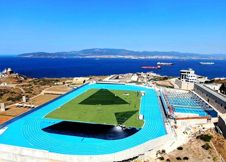 Aerial view of a coastal sports complex with a bright blue running track and pool overlooking the ocean.