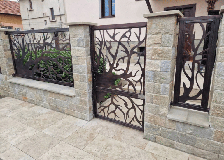 Modern wrought iron garden gate with an artistic tree branch design set in a natural stone fence.