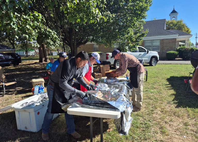 Volunteers at a community outdoor BBQ event preparing food on tables covered in aluminum foil.