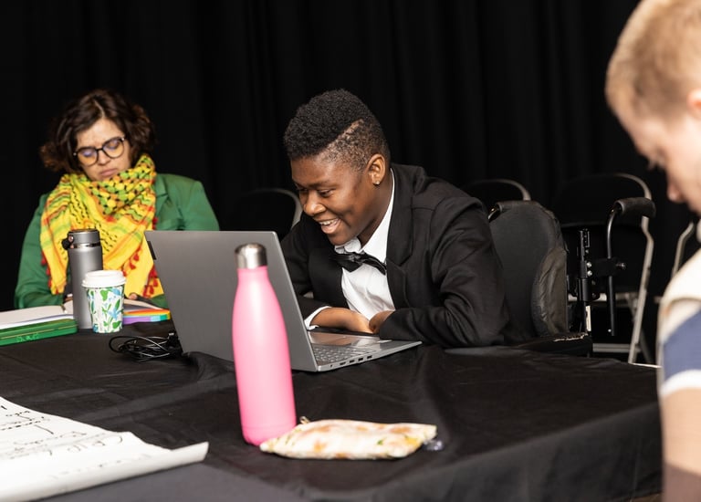 A mixed-race person in green, and black woman in a manual wheelchair sit at a table