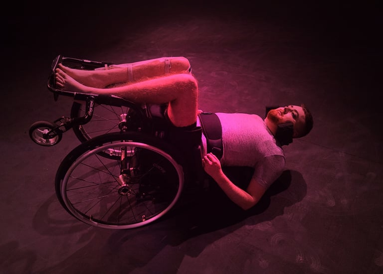 A half-dressed white person in a manual wheelchair on their back on the floor, bathed in pink light.