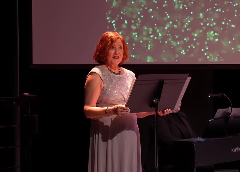 A female narrator on stage in a formal gown along with a music stand & piano.