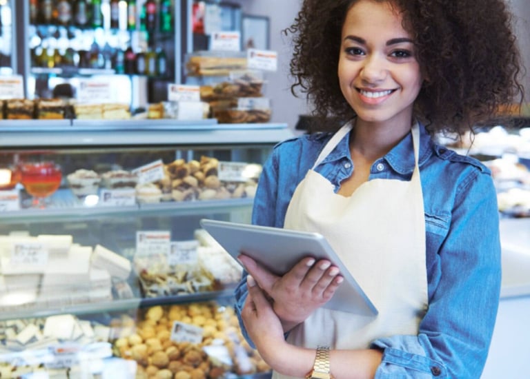 Pessoa em pé  com uma tablet na mão e no fundo uma vitrine de padaria