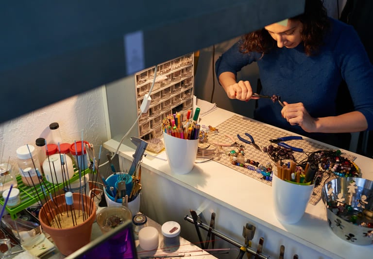 Jewelry maker using pliers at a desk filled with beads and artisan craft supplies.
