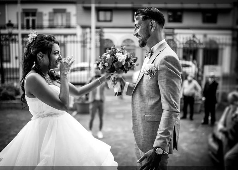 Mariée émue se couvre la bouche face au marié tenant le bouquet, N&B rue.