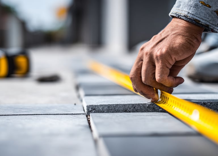 A contractor uses a yellow measuring tape to align grey stone pavers for a patio installation.
