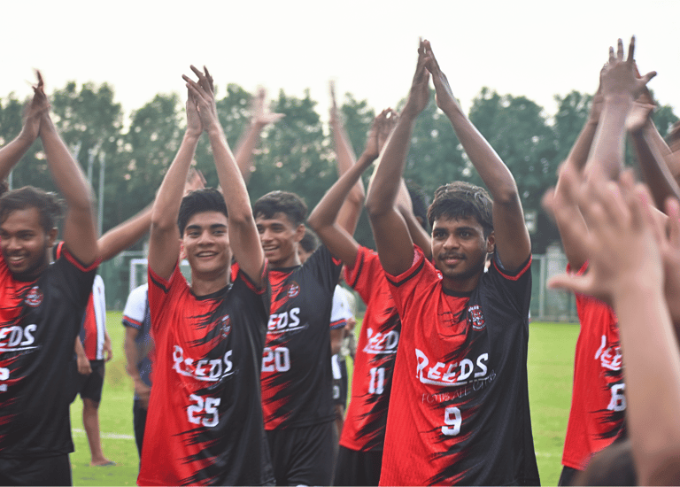 Reeds football club players in red and black jerseys clapping on a grass soccer field.