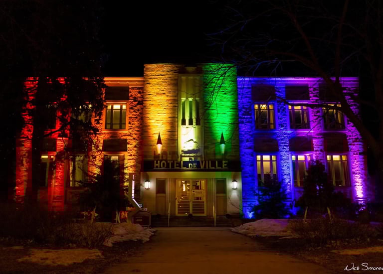 The Hotel de Ville building illuminated with vibrant rainbow pride colors at night.