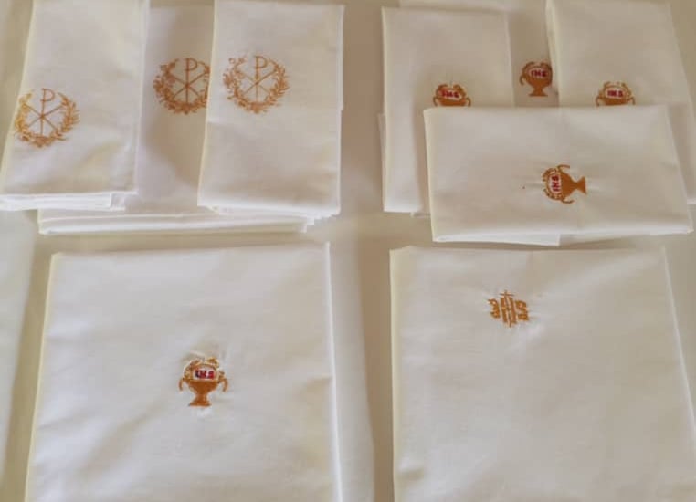 Stacks of white linen altar cloths featuring gold embroidered religious symbols like the IHS and Chi Rho.