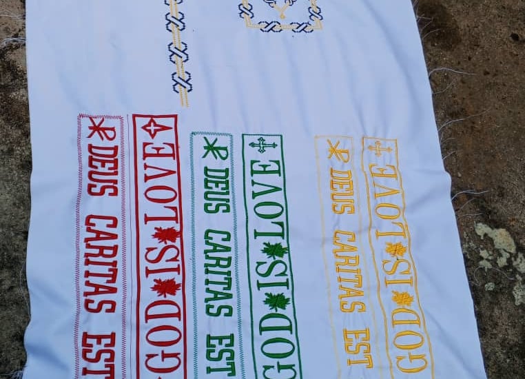 Hand-embroidered white linen featuring Deus Caritas Est and God is Love in colorful cross-stitch patterns.