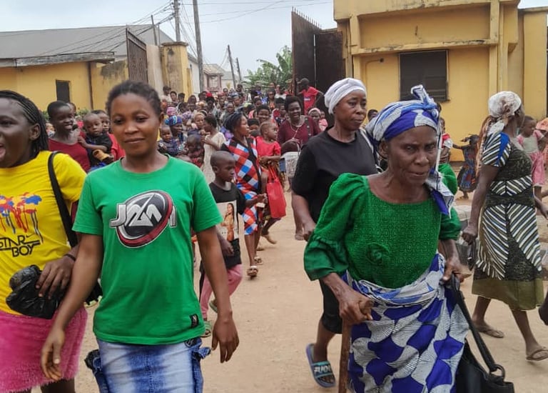 A diverse crowd of people, including an elderly woman with a walking stick, walking down a village street.