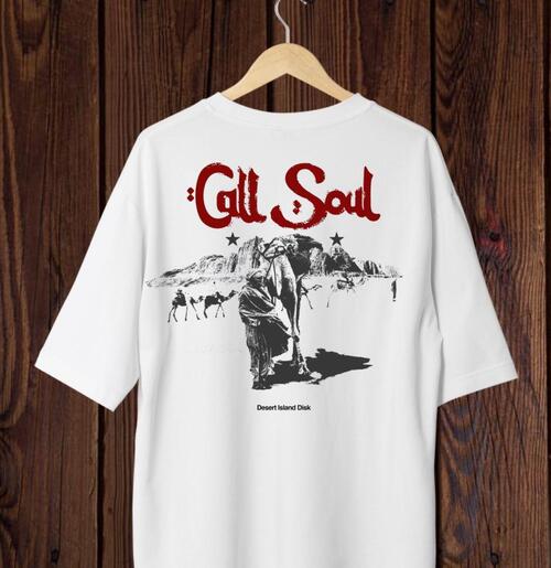 Camisa blanca con gráfico “Call Soul” y estampado de camello del desierto, colgada en pared madera.