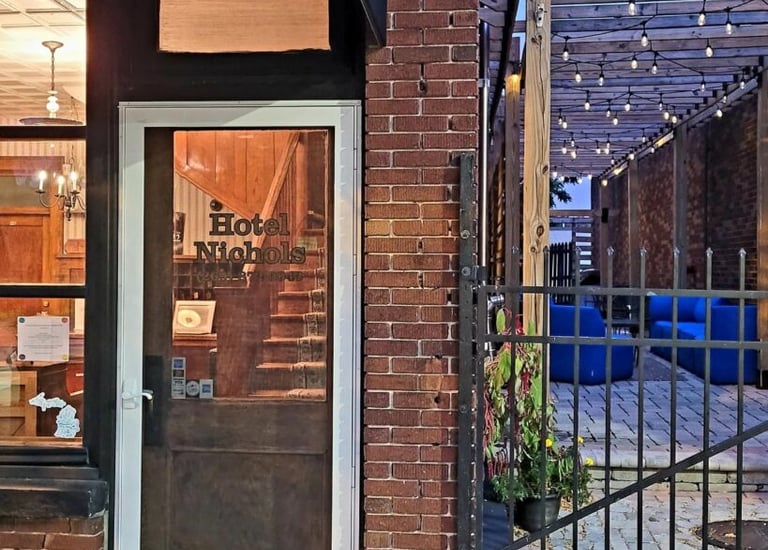 Entrance of Hotel Nichols with a glass door, brick facade, and adjacent patio area with string lights.