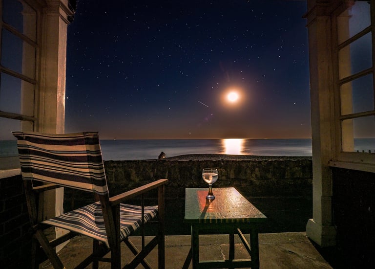 view of the moon over the sea as seen from a beach hut. A deck chair and glass on a table too