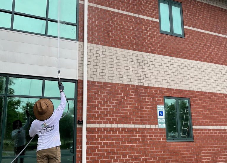 Professional soft wash technician cleaning a commercial brick building and windows with a pressure washer.